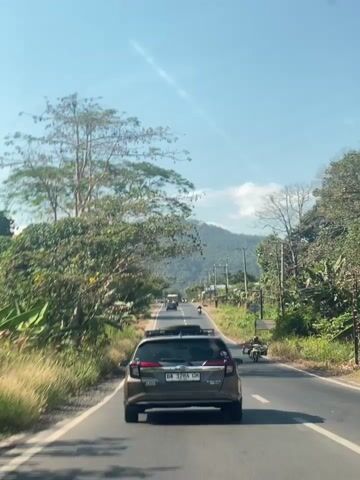 A point-of-view shot shows a vehicle driving down a paved road on a sunny day. The road is bordered by lush green vegetation and trees. In the distance, a mountain range can be seen under a clear blue sky. Several other vehicles, including cars and motorcycles, are also on the road, moving in the same direction as the viewer. A single motorcycle is visible on the right side of the road, passing the viewer's vehicle. The overall scene suggests a rural or semi-rural setting with active traffic.