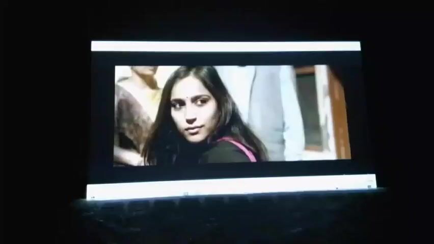 The video displays a scene projected onto a screen in a dark environment. The projected image shows a man dressed in a white outfit and a dark jacket walking towards a white vehicle. The man's face is visible. He appears to be entering or interacting with the vehicle. The scene outside the vehicle seems to be a street or an outdoor setting with some architectural elements. The video captures the man's movement and interaction with the vehicle in a short sequence.