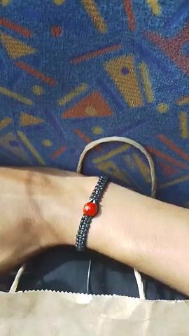 Here is a brief description of the video:

The video shows a person's wrist wearing a black, braided bracelet with a single red bead in the center. The bracelet is fairly tight on the wrist. The background features a blue surface with a pattern of abstract shapes in orange, yellow, and brown. A tan-colored bag strap is also visible in the frame.