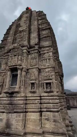 The video pans across the exterior of an ancient stone temple. The temple is ornately carved with intricate patterns and figures. The camera focuses on specific niches within the temple facade, each containing a small stone statue of a deity. The sky above is cloudy, and the overall impression is one of ancient grandeur and religious significance.