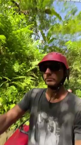 A man wearing a red helmet and sunglasses rides a red ATV through a lush, green forest. He is part of a group of people on ATVs, and they are driving on a dirt path with dense vegetation on either side. The camera is mounted to the ATV, providing a first-person perspective of the ride. The environment is bright and sunny.