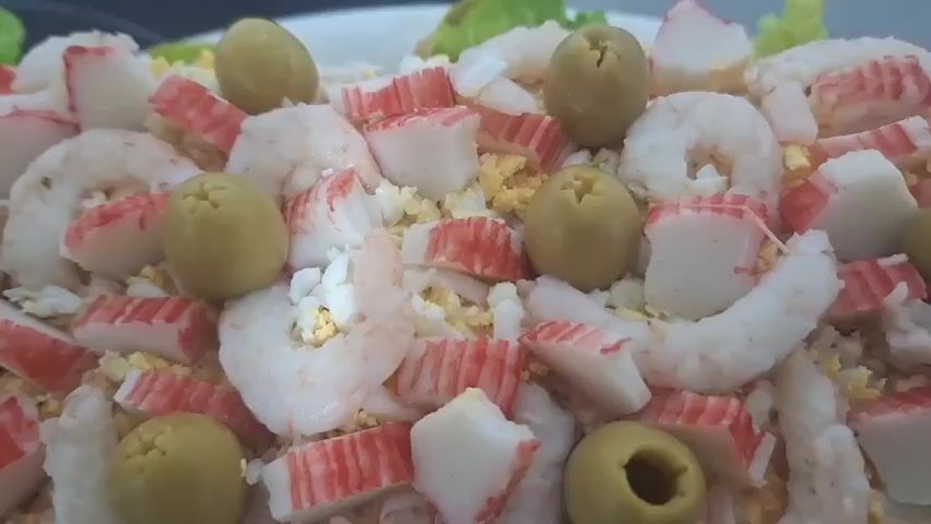 A close-up shot reveals a layered seafood dish, adorned with shrimp, crab sticks, hard-boiled eggs, and green olives. The dish is presented on a white plate, garnished with lettuce leaves. To the side, a separate plate holds a generous serving of cooked, unpeeled shrimp. The setting appears to be a kitchen, with a gray countertop visible beneath the plates.