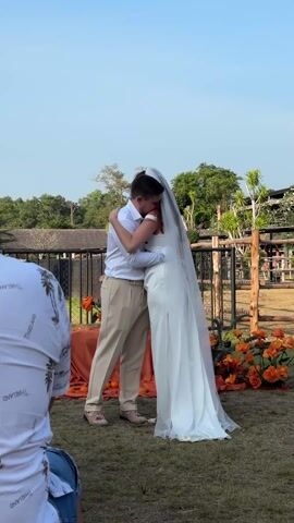 A wedding ceremony outdoors features a bride and groom in the center. The groom, wearing a white button-down shirt and khaki pants, embraces the bride, who is in a white gown with a long veil. They kiss amidst orange flowers and a matching orange table cloth on a table behind them. To the left, a man wearing a shirt with "Thailand" printed on it sits, possibly a guest or photographer. The backdrop includes a fence, greenery, and a blue sky. Cheers and applause can be heard.
