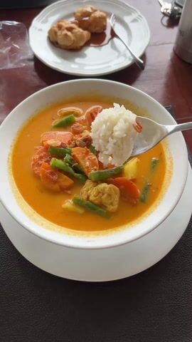 The video shows a bowl of orange curry with green beans, carrots, and tofu, topped with a scoop of rice. A spoon is holding the rice above the bowl. In the background is a white plate with what appears to be dessert. A set of keys and a thermos are also visible on the wooden table. The bowl sits on a white plate on a dark surface.