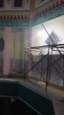 The camera pans upward in a room with a unique architectural design. The room features a green, elevated platform surrounded by walls painted in a combination of light yellow, white, and gray, accented with pink columns. Decorative Islamic calligraphy is visible near the ceiling, beneath a row of small, dark windows. Scaffolding stands against one wall, suggesting ongoing construction or renovation.  The room appears quiet and empty.