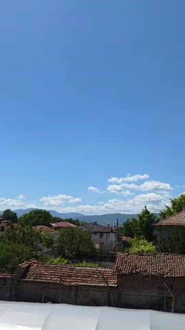 The video shows a view from a slightly elevated position looking out over a small town with traditional architecture. The rooftops are mainly red tile and the houses are one or two stories high. Green trees are interspersed among the buildings, partially obscuring some houses. In the distance, a mountain range is visible under a bright blue sky dotted with fluffy white clouds. The foreground shows a white, possibly tarp-covered structure, suggesting the viewpoint is from a building's rooftop or upper level. There is minimal movement, primarily the natural shifting of the clouds in the sky.