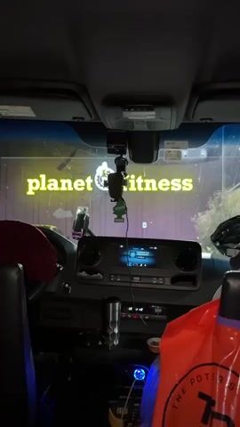 The video shows the interior of a vehicle, likely a van or truck, looking out through the windshield. The exterior is dark, and a bright yellow neon sign that says "planet fitness" is visible. A dashcam is mounted on the windshield above the rearview mirror. Below the dashcam, a car air freshener hangs from the mirror. The vehicle's dashboard is visible, with a digital screen showing what appears to be a music player or navigation system. There are also various accessories on the dashboard, including a phone mount with a phone. A red fabric is draped over the driver's side, and an orange bag is on the passenger side. The scene suggests the vehicle is parked outside a Planet Fitness gym at night.