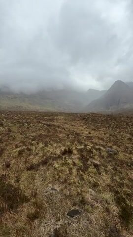 The video shows a wide shot of a mountainous landscape under a cloudy, overcast sky. The foreground is a barren, grassy field, tinged with brown and yellow, and dotted with a few rocks.  In the distance, a mountain range rises up, partially obscured by a heavy layer of clouds and fog that hangs low.  The overall impression is one of bleakness and isolation. There are no people visible in the frame.