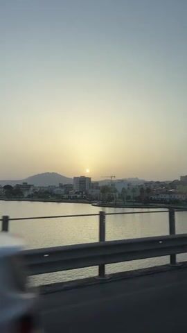 The video shows a view of a coastal city at sunset. The sun is low in the sky, casting a warm glow over the scene. Buildings line the waterfront, and palm trees dot the landscape. A crane is visible in the background, suggesting ongoing construction. The video appears to be filmed from a moving vehicle, possibly a car or bus, as evidenced by the blurred foreground of a guardrail and road surface. The water in the foreground reflects the sky and the city lights. The overall atmosphere is peaceful and serene.