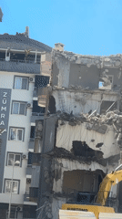 Excavator demolishes damaged multistory building, debris scattered throughout area
