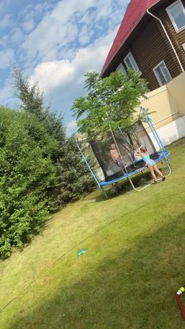 Two children are jumping on a trampoline in a grassy backyard. A young boy in striped shorts is on the left side of the trampoline, and a young girl in a light blue swimsuit is on the right. The girl appears to be pushing off the trampoline mat with her hands. A white dog is lying down near the boy. A large wooden house with a red roof is visible in the background, along with a beige fence and some trees. The sky is blue with scattered white clouds.