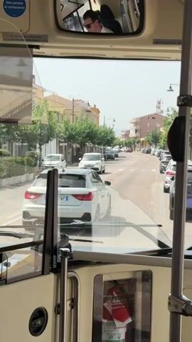 The video captures a view from inside a bus looking out the windshield onto a street scene. In the foreground, the dashboard and a fire extinguisher are visible. The bus is moving slowly through a town. On the street, several cars are driving or parked. A white Audi A1 is the most prominent car, seen from the rear, as it drives ahead of the bus. Trees line the street, with buildings on either side. The sky is bright and clear. The video appears to be filmed during the daytime.