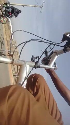 The video shows a person riding a bicycle on a paved road under a clear, bright sky. The person is wearing a striped polo shirt and brown pants, and they are holding the handlebars. A building with a pyramid-shaped roof is visible in the background. The camera appears to be mounted on the bicycle, providing a first-person perspective of the ride.