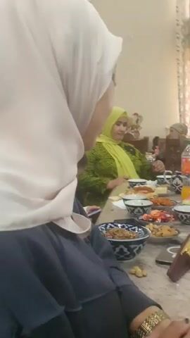 The video shows a gathering of people, likely a family, seated around a table laden with food. Several women are visible, some wearing hijabs. A young girl is looking at her phone. There are various dishes of food, including what appears to be bread, nuts, and fruit. One woman in the foreground is looking at the camera and makes a hand gesture. The environment appears to be indoors, possibly a dining room, with curtains visible in the background. The overall atmosphere seems casual and celebratory.