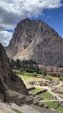 The video captures an aerial view of an ancient Inca site with a large mountain in the background. In the foreground, there are stone terraces and ruins. Beyond the ruins, there is a green grassy field with some structures. Further in the distance, a town with red-tiled roofs is visible, nestled between the mountains. People are scattered throughout the ruins and town, with some walking on the terraces and others in the town. The sky is blue with scattered white clouds.