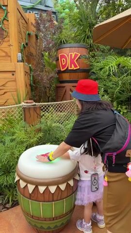In a vibrant theme park setting, a woman in a red and blue bucket hat guides a young child's hand to hit a large, interactive white and brown drum. The drum emits a pink glow where it's touched, with red cards or items visible on its surface. In the background, other drums of similar design are seen, and a crowd of adults and children observe the surroundings.

The area is richly themed with jungle-like foliage, wooden structures, and rock formations featuring wooden spikes. A prominent wooden barrel with "DK" (Donkey Kong) written on it is visible, confirming the setting as part of the Donkey Kong Country area within Super Nintendo World, Universal Studios Japan. A wooden post with hanging bells adds to the immersive environment.

The weather appears bright and clear. The general atmosphere is lively and joyful, with people enjoying the interactive elements and attractions of the park.