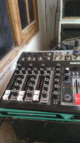 The video shows a close-up shot of a Bomge KV4 audio mixer. The mixer is black with silver and red accents. It has multiple knobs, sliders, and input jacks. A blue and black cable is plugged into the top left of the mixer. The mixer is placed on a wooden surface next to a window frame with a glass pane. In the background, a speaker grille is visible. The lighting appears to be natural daylight. The camera is slightly tilted and pans slowly across the mixer from left to right.