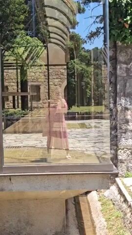 This video captures a reflective scene with a woman in a pink and white patterned dress walking in front of a glass-enclosed structure. The glass acts as a mirror, reflecting the surrounding environment. We see a stone building with arched windows and a tiled roof, as well as lush greenery and trees, suggesting a sunny day. The ground is paved with stones, and there's a small channel of water running along the base of the glass structure. To the right of the glass, there's a stone wall with an arched entrance and a gray wooden door. Beyond the immediate area, more trees and a glimpse of a grassy field under a bright blue sky are visible. A green trash bin is also present in the scene, adding an everyday element. The overall impression is one of a peaceful, possibly historic, outdoor setting in Cetinje, Montenegro, during daylight hours with clear weather. The woman's reflection is the main human subject, appearing to be strolling through this picturesque location.