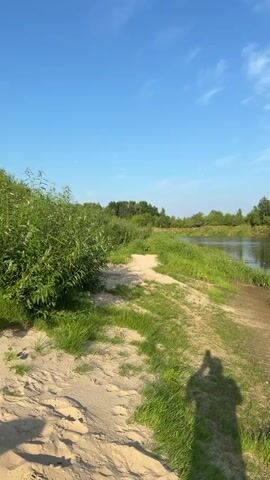 The video captures a serene river scene on a bright, sunny day. A sandy bank with green grass lines the edge of a calm, reflective river. Lush green trees and vegetation form a dense backdrop on the opposite bank, their reflections shimmering on the water's surface. The sky above is a vibrant blue, dotted with wispy white clouds. In the foreground, a shadow of a person, likely the videographer, is cast on the sandy path, indicating their presence. The overall atmosphere is peaceful and natural, suggesting a tranquil outdoor setting.