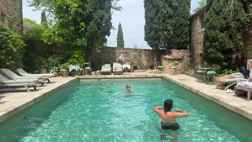 This video captures an outdoor swimming pool area, likely at a private residence or villa in Saint-Victor-la-Coste, France, bathed in bright daylight. The rectangular pool, filled with clear blue-green water, is the central focus.

Two individuals are in the pool: one person is treading water near the center, and another is closer to the foreground, facing away from the camera, with their back to the viewer. On the right side of the pool, a person is relaxing on a lounge chair, dressed in light-colored clothing.

The pool area is surrounded by rustic stone walls covered in lush green foliage, suggesting a historical or traditional architectural style. Tall, slender cypress trees punctuate the landscape, adding to the Mediterranean aesthetic. Several lounge chairs are arranged around the pool, along with small tables and chairs, indicating areas for relaxation and socializing. The overall atmosphere is one of leisure and tranquility.