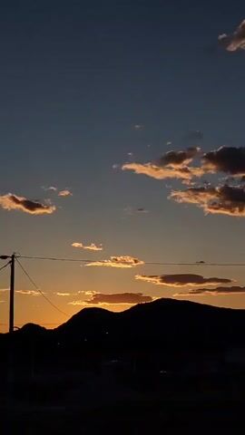 This video showcases a serene twilight or dusk scene in Mahala, Montenegro. The sky is a deep blue-grey, transitioning from lighter blues to darker tones, with scattered clouds that catch the last light of the setting sun, revealing hints of orange, pink, and light grey.

The foreground elements are silhouetted against the sky, including distant mountains or hills, various trees with dense foliage, power lines spanning across the frame, a street lamp, and a white building with visible windows.

The camera pans slowly, capturing different compositions of the sky and the dark landscape below. The overall atmosphere is peaceful and calm, typical of late evening. No people or specific activities are visible, and there is no discernible text.