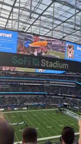 The video shows a football game at SoFi Stadium in Los Angeles, CA. The massive video screen displays highlights and promotional material. The stands are filled with fans, and the football field is visible below.