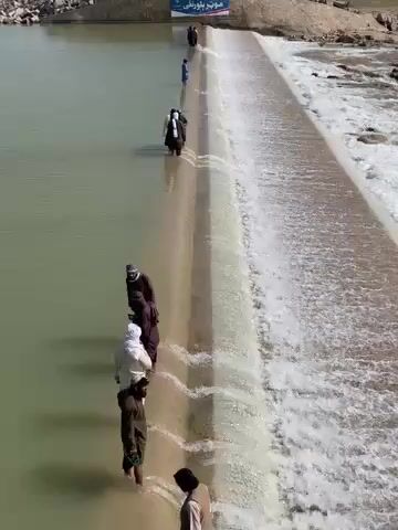 The video captures an overhead shot of a group of people walking across a low dam or weir. The concrete structure spans a body of water. On the left side of the dam, the water is still and appears murky green, while on the right side, water cascades over the dam, creating a white, foamy waterfall.

Approximately a dozen individuals, primarily men, are visible walking in single file across the top of the dam. Their lower legs are submerged in the water covering the dam's surface on the upstream side. Many appear to be barefoot or wearing sandals. They are dressed in traditional loose-fitting clothing, including long robes and tunics, in various shades of brown, black, and white.

One person in the foreground, wearing dark clothing, is walking carefully. Further back, another individual in white clothing follows. In the mid-ground, a person in dark clothing is seen carrying a child on their shoulders. The people are moving from left to right across the structure, seemingly using it as a pedestrian crossing.

The surrounding landscape appears arid, with sandy-brown banks visible on either side of the water. In the distant background, on the left bank, a blue sign with white text, possibly in an Arabic-script language, is visible, but the words are unreadable. The scene is brightly lit by natural sunlight, suggesting it is daytime. The sound of rushing water from the cascading dam is clearly audible throughout the video.