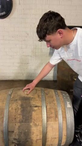 A young man is shown inspecting a wooden barrel. The man has short brown hair, and is wearing a white T-shirt and gray pants. He is standing in a room with a brick wall and a concrete floor. The barrel is lying on its side, and there is a small hole in the top. The man places his mouth near the hole, appearing to sniff the barrel. Then he puts the cork back in the hole, smiles, and steps away from the barrel. Another barrel and a wooden box are in the background.