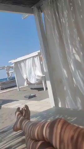 The video shows a first-person perspective of someone relaxing on a beach. In the foreground, bare feet are visible, casting shadows on the sand. In the midground, there are several white beach cabanas with draped white curtains. A woman in a colorful sarong and bikini walks from left to right across the sand towards the foreground. She pauses and turns to face the camera, spreading her arms wide. Another person is visible in a cabana on the far left. The sky is clear and blue.