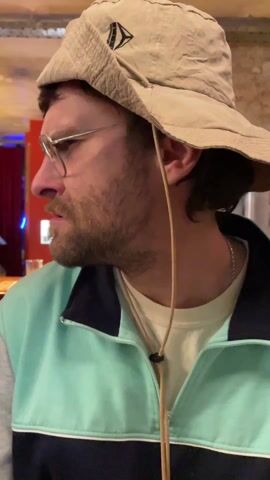 The video shows a man wearing a beige bucket hat, wire-rimmed glasses, and a light blue track jacket with a black collar. He has a mustache and a short beard. He looks to his left, then directly at the camera, smiling slightly. He then looks to his right and back to his left again. He appears to be indoors, possibly in a bar or restaurant setting. There is a red curtain and some blurred lighting in the background. The camera angle is close-up.