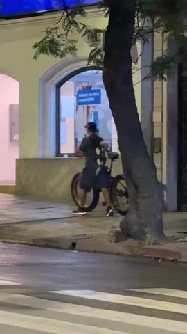 The video shows a man walking with a bicycle next to him. He is wearing a dark shirt, dark shorts, black shoes, and a baseball cap. On his back, he is carrying what appears to be several small stuffed animals. He walks along the sidewalk past a building with an arched entrance and windows. There are people walking on the sidewalk, trash cans, and a parking sign that says "EXCLUSIVO." There are also trees in the scene. The video appears to be taken at dusk or night, with limited lighting.