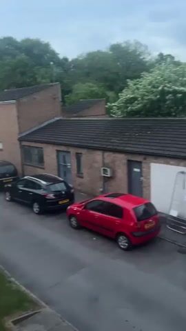 The video shows an outdoor view of a residential street lined with parked cars and brick buildings. The view is initially from a high angle, possibly a window, looking down at the street. The scene is somewhat dark, indicating it could be dusk or early evening. The street is wet, suggesting it might have rained recently.  Trees are visible in the background, adding to the residential setting.  The video then focuses on a window being opened, with a hand visible holding the window frame. The focus shifts to the window itself, reflecting the surrounding buildings and sky.