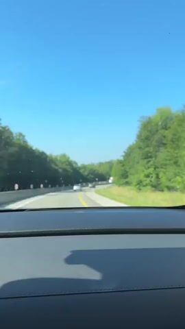 A first-person perspective video shows a view from the inside of a car driving on a highway. The sky is clear and blue, and lush green trees line both sides of the road. Several cars and a truck are visible on the road ahead. The car appears to be moving at a moderate speed, and the scenery is passing by smoothly. The interior of the car, including the dashboard, is visible in the foreground.