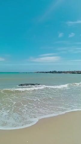 The video shows a beautiful beach with clear blue skies and calm waters. The sand is light brown and there are small waves rolling onto the shore. There is a large rock in the water, and some people are visible in the distance. The video pans across the beach, showing the expanse of sand and sea. In the background, there are some structures and hills. The overall atmosphere is serene and peaceful.