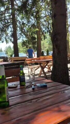 The video pans across an outdoor seating area in a wooded environment. In the foreground, a wooden table and bench are visible. On the table are a bottle of beer, an empty glass, a cigarette lighter, and a pack of cigarettes. In the background, several picnic tables with benches are set up on a gravel ground, surrounded by trees. People are seated at some of the tables, engaged in conversations or eating. Further in the background, there are cars parked and a small wooden structure that appears to be a bar or a kiosk. The overall impression is that of a relaxed outdoor restaurant or cafe.