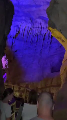 The video shows a group of people walking on a wooden walkway inside a cave. The walls of the cave are illuminated with blue, purple, and yellow lights. The people are walking in the same direction. One person in the group is holding a smartphone and taking a picture of the cave. The cave walls have jagged features.