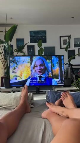 The video shows a couple lying in bed, watching a television. The television displays a scene from the Netflix series "Wednesday," featuring Jenna Ortega as the titular character. The room appears to be a bedroom, with a large television placed on a stand in front of the bed. Several large potted plants are situated around the room, adding a touch of greenery to the setting. The walls are adorned with various framed artworks, suggesting a decorative and personal touch to the space. The lighting indicates it is daytime, with natural light filtering into the room. The couple's feet are visible in the foreground, one person's feet are bare, and the other's are adorned with dark nail polish. The overall mood seems relaxed and cozy, as the couple enjoys a shared viewing experience. The visible text on the television screen is in Spanish and appears to be subtitles for the show. The image captures a moment of leisure and entertainment within a home environment.