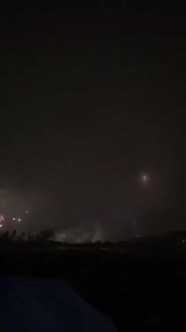 This video captures a nighttime scene with fireworks illuminating the dark sky. In the foreground, a dark silhouette of land is visible, with some trees. In the middle ground, multiple bursts of fireworks explode, creating bright flashes of light and smoke. The fireworks appear to be launched from behind a line of trees. In the background, the sky is dark, with some faint light sources visible in the distance. The overall atmosphere is festive and celebratory.