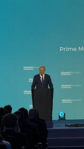 A man in a dark suit and tie stands behind a black podium on a stage. He is speaking, with a serious expression on his face. The background is a large screen with a repeating logo that says "Ukraine Recovery Conference" and "Prime". There are audience members in the foreground, silhouetted against the stage lighting.