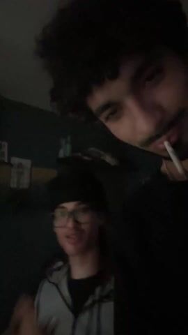 Two young men appear in a low-lit room, recording themselves with a front-facing camera. One man, with dark curly hair and a mustache, holds the camera close to his face. The other, wearing glasses and a black head covering, stands slightly behind. Music plays in the background as the man with glasses speaks and gestures. A guitar is partially visible in the background, along with some other items on a shelf.
