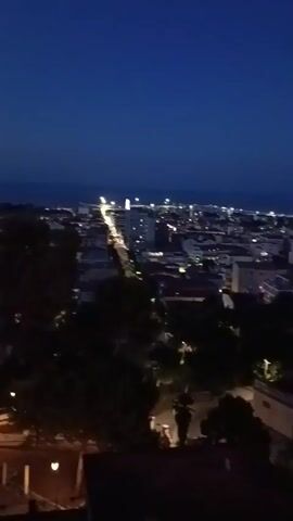 This media file captures a nighttime view of Giulianova, Italy, looking out over a town illuminated by artificial lights. The scene is set against a deep blue twilight sky.

The foreground shows a dark, paved area, possibly a terrace or plaza, with some low-lying greenery. A few blurry figures can be seen seated or standing in this area, suggesting a relaxed atmosphere. Further into the frame, the town sprawls below, a dense collection of buildings dotted with countless lights. Streets are visible, marked by rows of streetlights, and the overall impression is of a vibrant, inhabited place at night.

The time of day is clearly evening or night, given the dark sky and the prevalence of artificial lighting. The weather appears clear, with no signs of rain or strong winds. The architecture of the buildings suggests a European setting, consistent with the provided location context of Giulianova, Italy. The overall mood conveyed is one of peacefulness and the gentle hum of a town settling into the night. There are no discernible text elements within the visible frames.