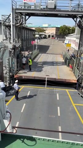 The video shows a view from the deck of a ferry looking towards the ramp and the shore. Two uniformed individuals are visible on the ferry deck in the foreground, one facing towards the ramp and the other towards the shore. The ramp is lowered and appears to be made of metal, with yellow lines indicating lanes. On the shore side, beyond the ramp, there are railings and a building with windows. A few people are visible on the shore, including one person wearing a yellow safety vest. The scene appears to be a ferry terminal on a sunny day.