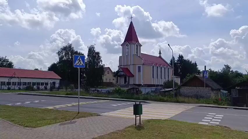 The video shows a street scene with a church in the background. A two-story building with a red roof is visible on the left. A church with a red steeple and white and yellow walls is in the center. There are trees and a few houses on the right. A pedestrian crossing is marked on the road in the foreground, and a green trash can is next to it. A blue pedestrian crossing sign is on a pole to the left of the crossing. The sky is partly cloudy.