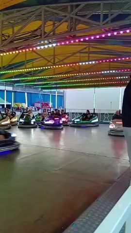 The video shows a bumper car ride at an amusement park. Several people are riding in colorful bumper cars. The cars are moving in different directions and bumping into each other. The ride is illuminated by strings of colorful lights that run along the ceiling. The ground is a dark, smooth surface, possibly concrete or asphalt. In the background, there are tents and other structures, suggesting an outdoor event or fair. The overall atmosphere appears to be fun and lively.