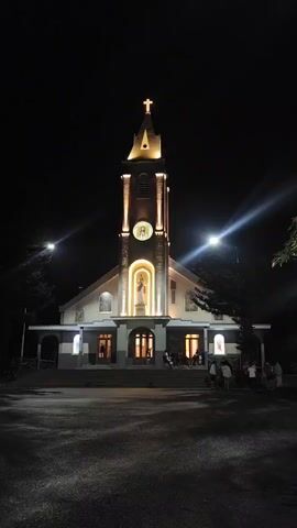 The video displays a church at night, illuminated by external lights. The camera is positioned low, looking up at the church facade, which is lit to highlight architectural features. A cross is visible atop the tallest spire, also illuminated. Below the cross, a clock face is depicted, and further down, a statue of a religious figure stands within an arched niche, bathed in warm light. The entrance of the church is visible, with several doors and a set of stairs leading up to it. A few people are gathered on the steps and near the entrance, appearing to be socializing. The surrounding environment is dark, suggesting it is nighttime, with some trees visible on the right side of the frame. The overall impression is of a peaceful evening scene at a place of worship.