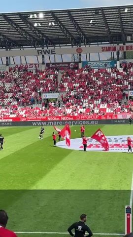 The video shows a soccer stadium on a sunny day, packed with fans wearing red clothing in the stands. The field is bright green, with players visible on the pitch. In the foreground, a group of people in red tops and black pants are holding red and white flags, creating a visually coordinated display. The stadium is covered with a metal roof, and various advertising banners are visible along the sides. There is a person in black at the bottom of the screen, facing away from the camera, wearing what looks like a training jacket.