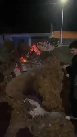 At night, the camera pans from a large pile of logs and glowing embers to a group of people standing in a paved plaza in front of a small church building decorated with strings of blue and white lights. A streetlamp provides additional illumination. One man wearing a dark jacket and jeans stands next to the burning logs. Another person in a white hooded sweatshirt walks past him. Two men and a boy stand in the plaza in front of the church. The church building has a small bell tower with orange figures illuminated inside. A large white star hangs above the entrance.