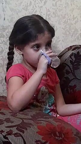 A young girl with dark hair in pigtails sits on a patterned sofa, drinking from a clear glass. She is wearing a pink t-shirt with a graphic of Elsa from Frozen on it and pink pants. Her left arm is bent, and her right arm is extended. The background is a textured, light-colored wall.