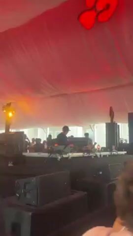 The video shows a music performance under a large white tent, illuminated with red lights. A DJ, wearing a cap, is mixing music on a stage crowded with equipment. A group of people can be seen watching the performance in the background. Large speakers are positioned in front of the stage, and some audience members are visible in the foreground.
