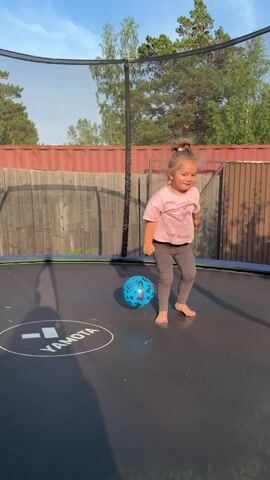 A young girl with blonde hair tied in a bun is playing on a trampoline. She is wearing a pink t-shirt and gray leggings. A blue ball with white circles is on the trampoline. The girl jumps and then sits down, and then crawls on the trampoline. The background shows a wooden fence and trees. The sun is shining.